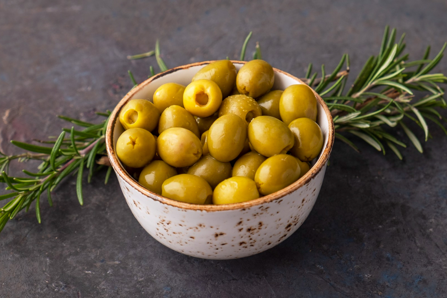 A glass cruet filled with golden extra virgin olive oil, surrounded by green and black olives and a sprig of rosemary.